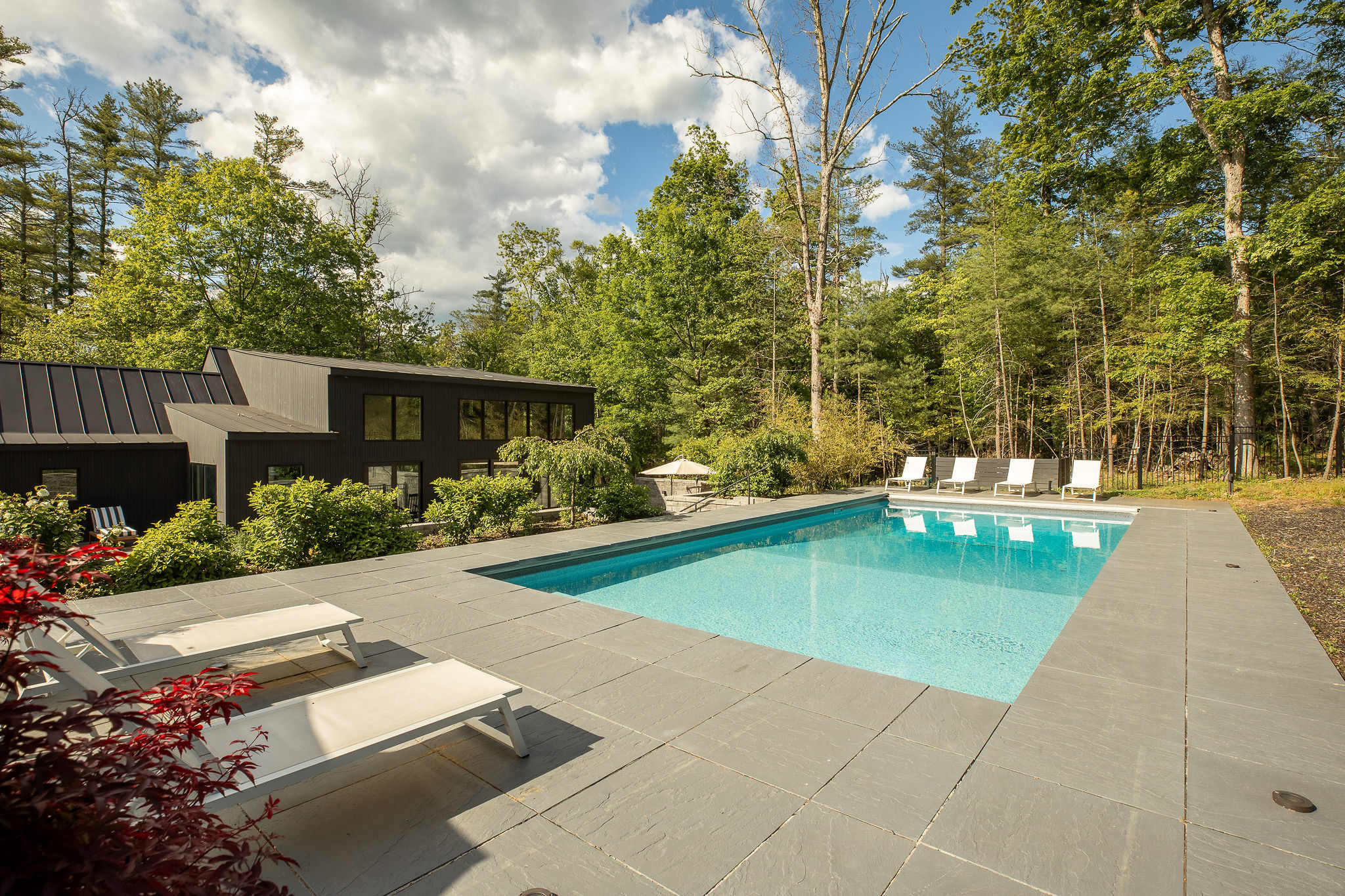 Inviting outdoor swimming pool with light blue water, surrounded by dark grey stone paving and white lounge chairs. The modern black house with large windows and lush trees provides a serene backdrop.