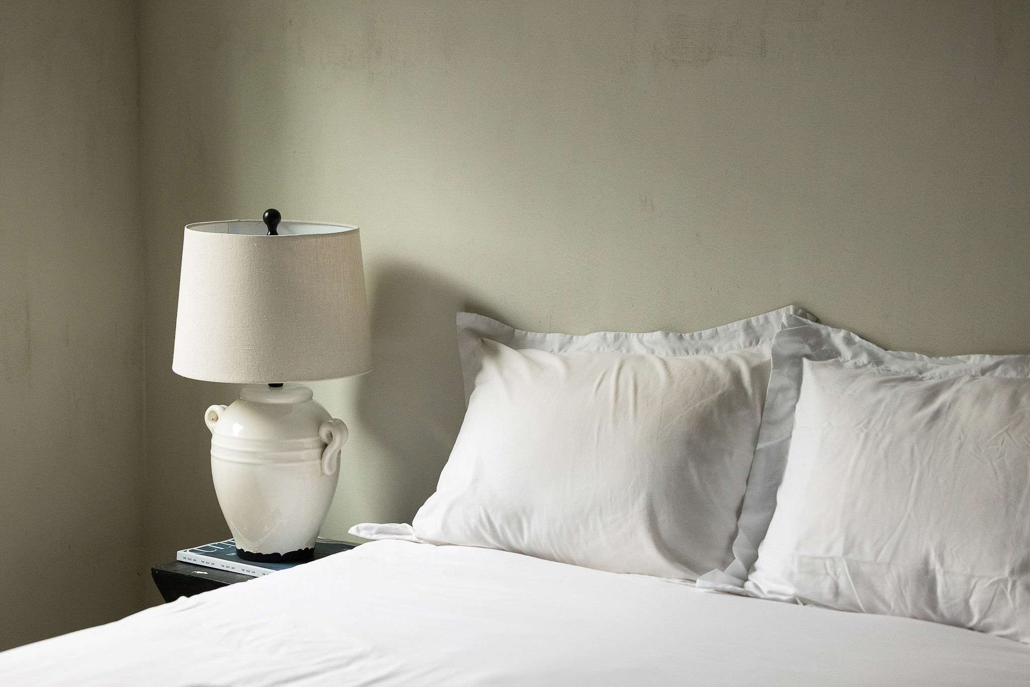 A close-up of a bed with crisp white pillows and bedding, next to a white ceramic lamp on a dark nightstand. The light-colored wall has a subtle texture.