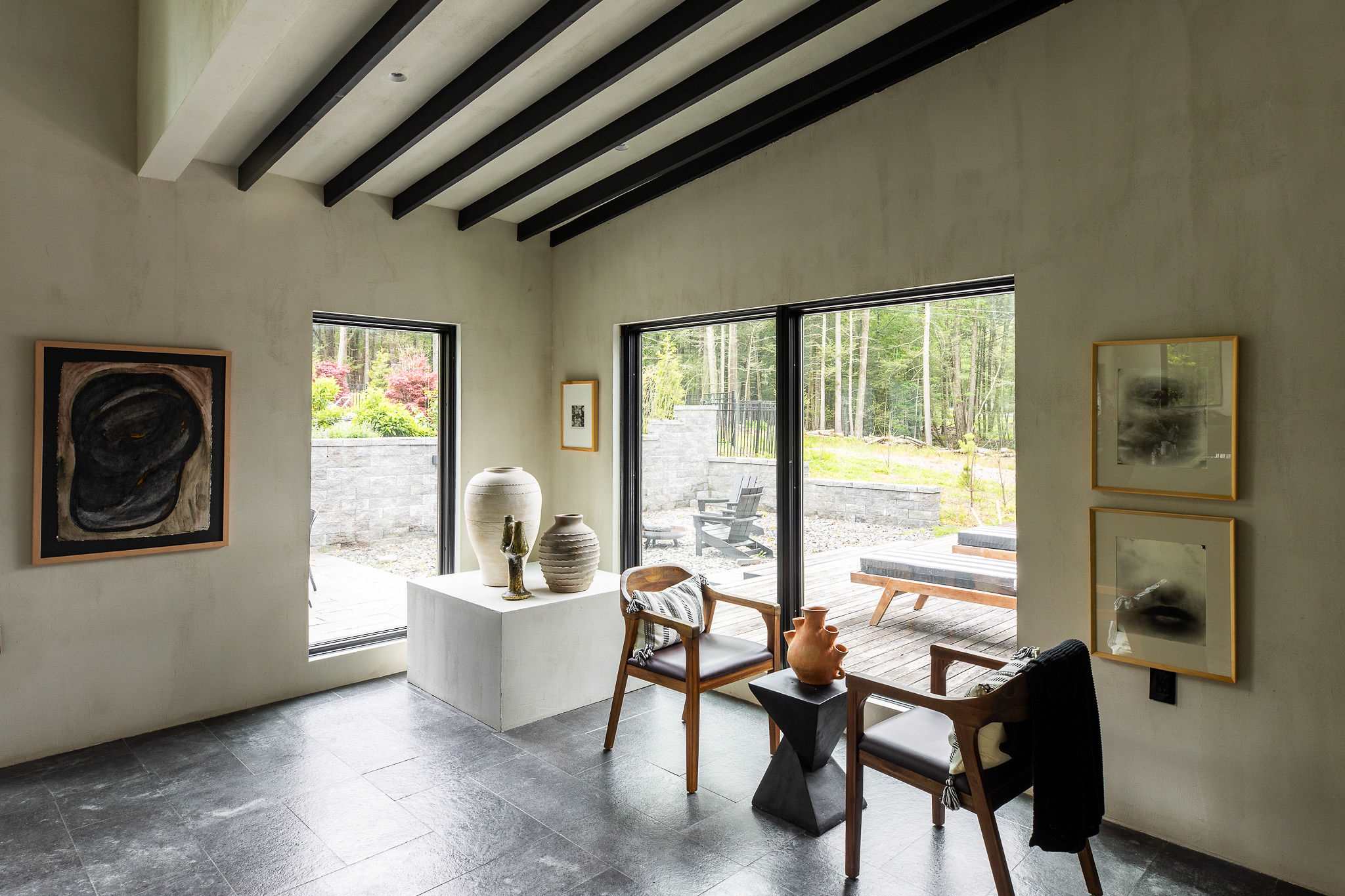 A bright living area with dark tiled floors and exposed black ceiling beams features two wooden chairs with leather seats, decorative vases on a white pedestal, and large windows overlooking an outdoor patio.