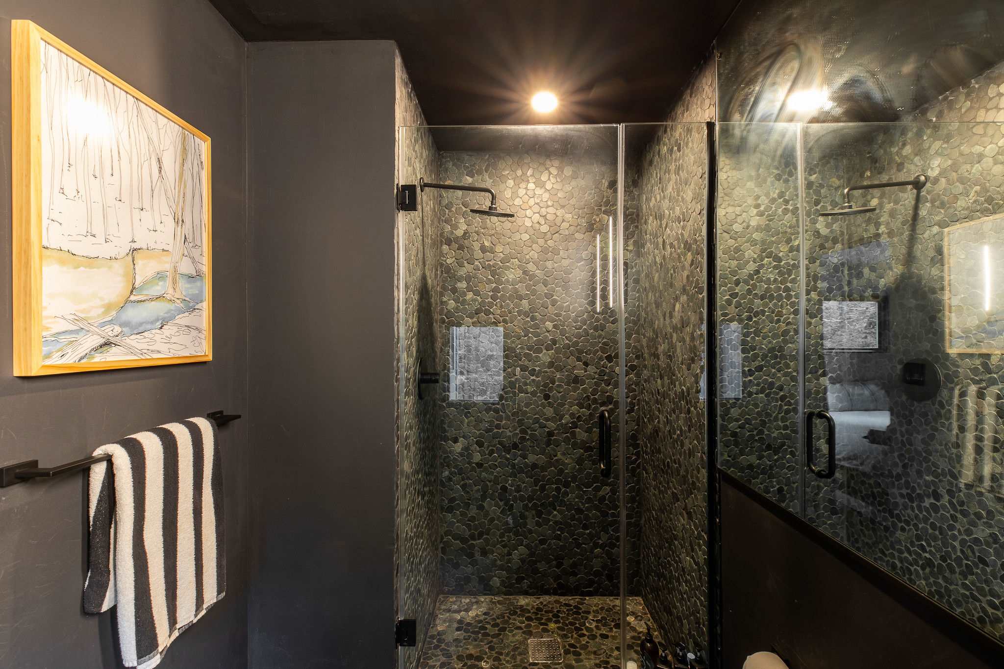 A dark-toned bathroom with a glass-enclosed shower featuring dark pebble-tiled walls. A framed artwork hangs on the dark gray wall, and a striped towel is draped over a black towel bar.