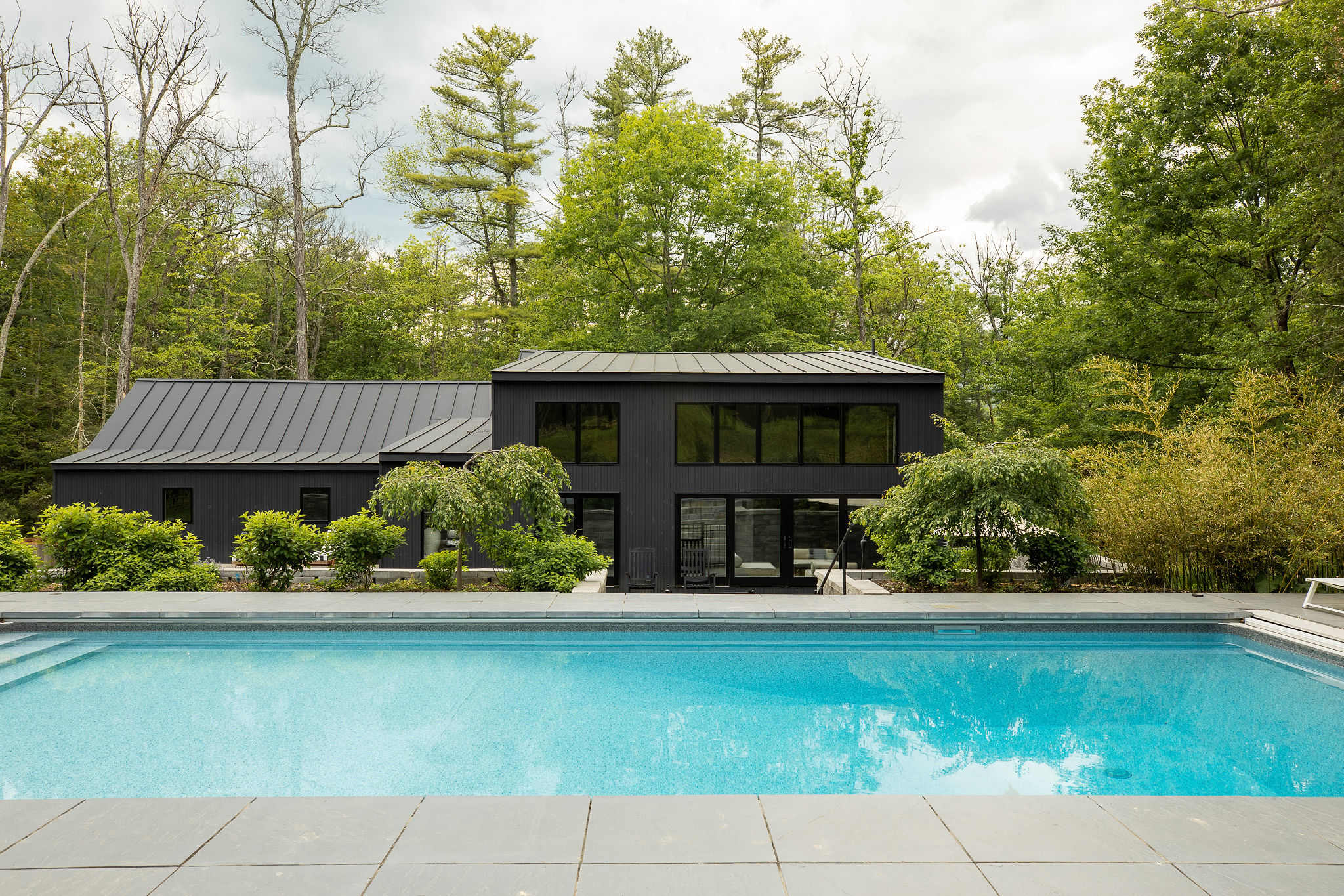 Wide shot of the modern black house with a dark metal roof, viewed from across the rectangular swimming pool. The pool area is surrounded by dark grey stone paving and lush green trees, creating a private oasis.
