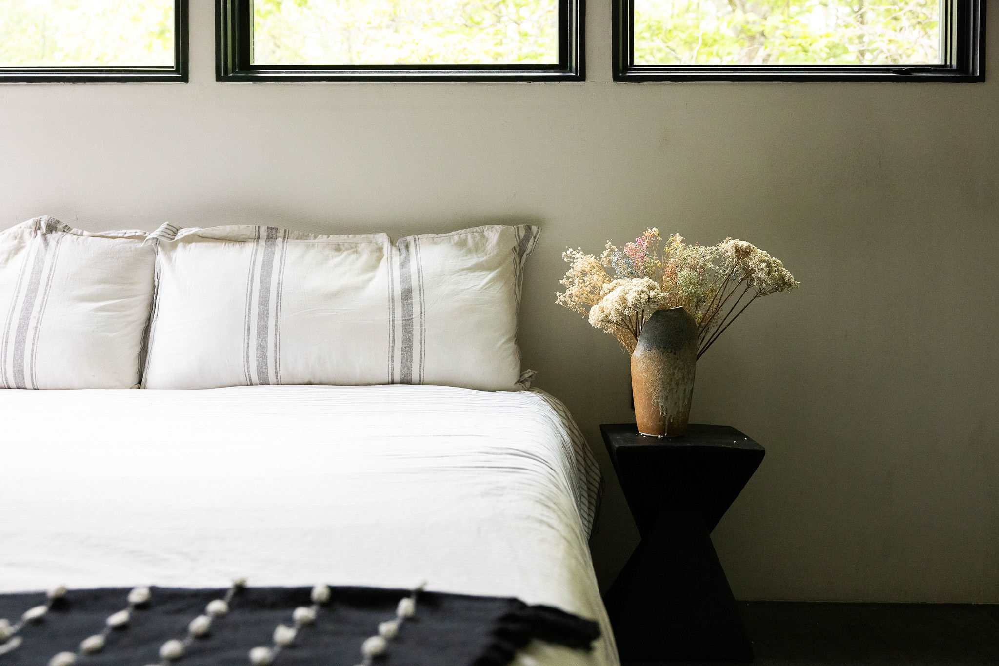 A close-up of a bed with white striped pillows and a white duvet, accented by a black and white patterned throw blanket. A small black nightstand holds a vase with dried flowers.