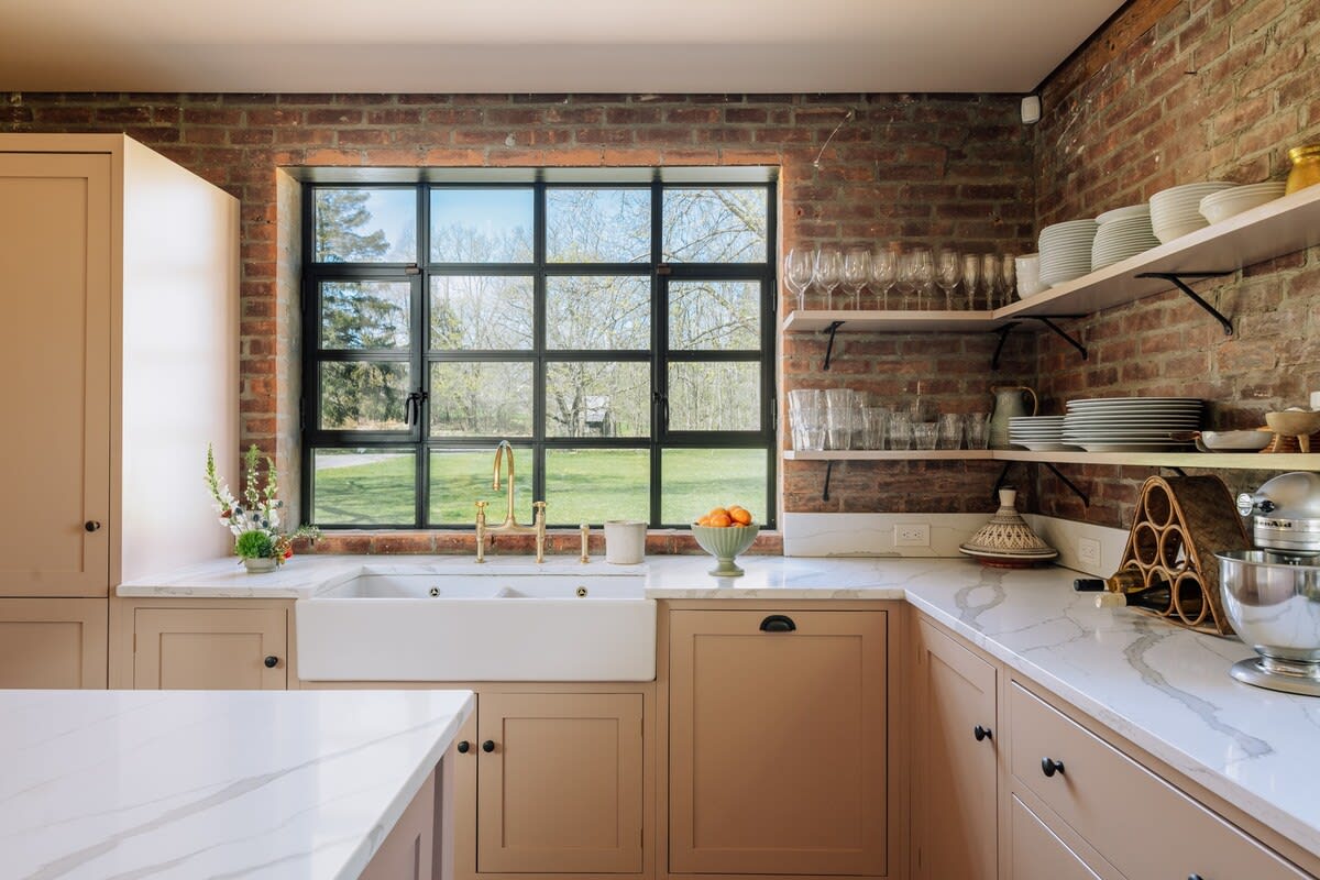 Bright kitchen corner with exposed brick walls, light brown cabinetry, white marble-patterned countertops, and a large farmhouse sink under a grid-paned window overlooking greenery.
