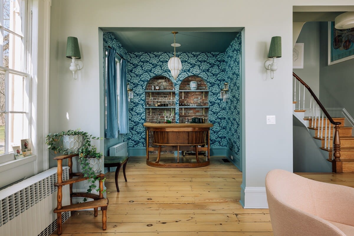 A view into a distinct bar alcove with blue patterned wallpaper and exposed brick shelves, seen from a light-colored room with hardwood floors and large windows. A wooden staircase with white risers is visible to the right.