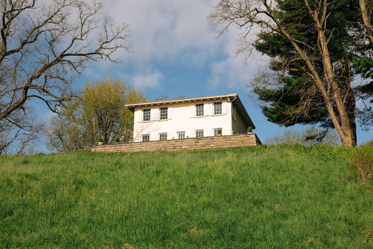 Ground-level view of a white two-story house with dark shutters, perched atop a lush green hill, framed by bare trees against a bright blue sky with scattered clouds.