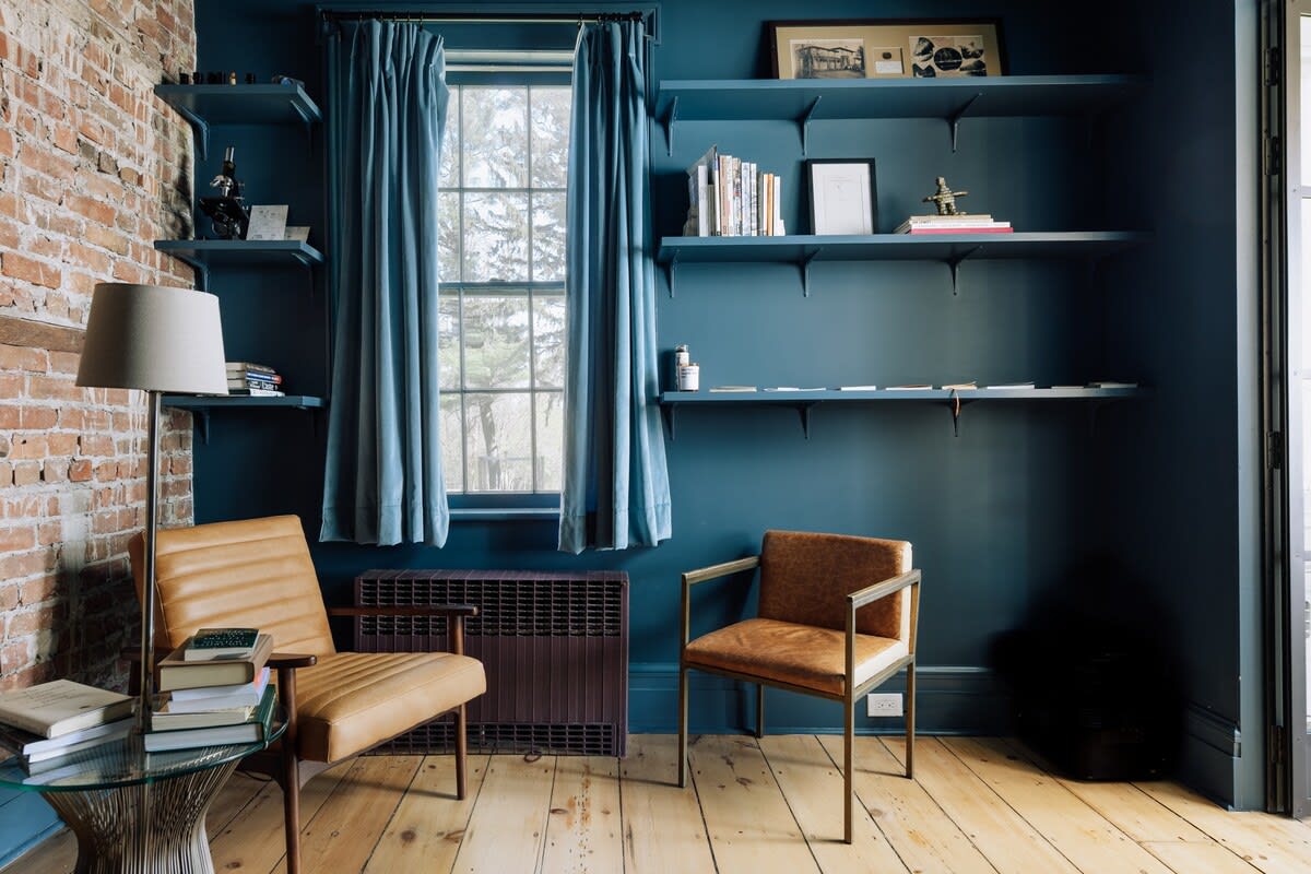 A cozy study or den with dark blue walls and built-in shelves, featuring a large window with blue curtains, and two modern leather armchairs. Exposed brick is visible on one wall, and books are neatly arranged on a glass side table.