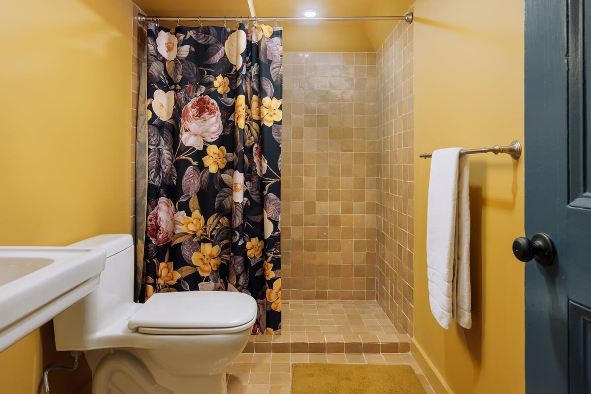 A vibrant bathroom with bright yellow walls and a tiled walk-in shower, featuring a bold floral shower curtain with dark and light flowers. A white toilet and a simple sink are also present.