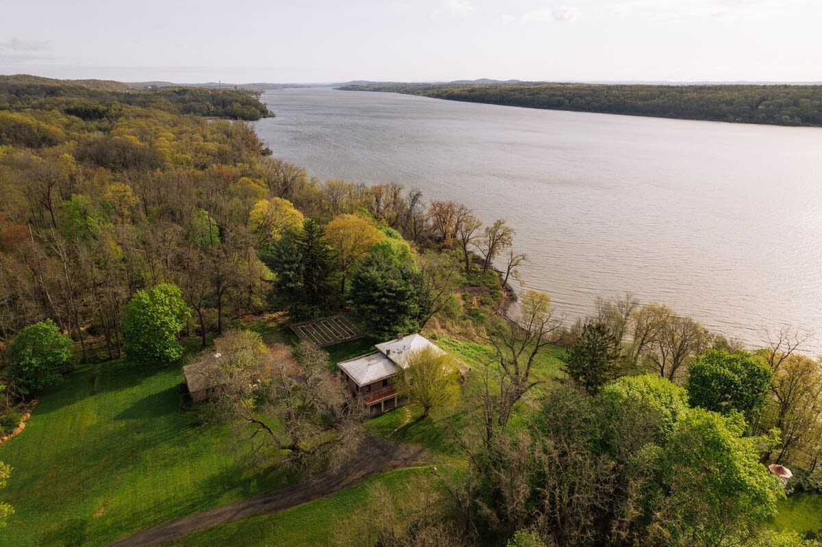 High aerial view showcasing the main house and a smaller building nestled on a green hillside, surrounded by lush trees, with a wide river flowing alongside the property.