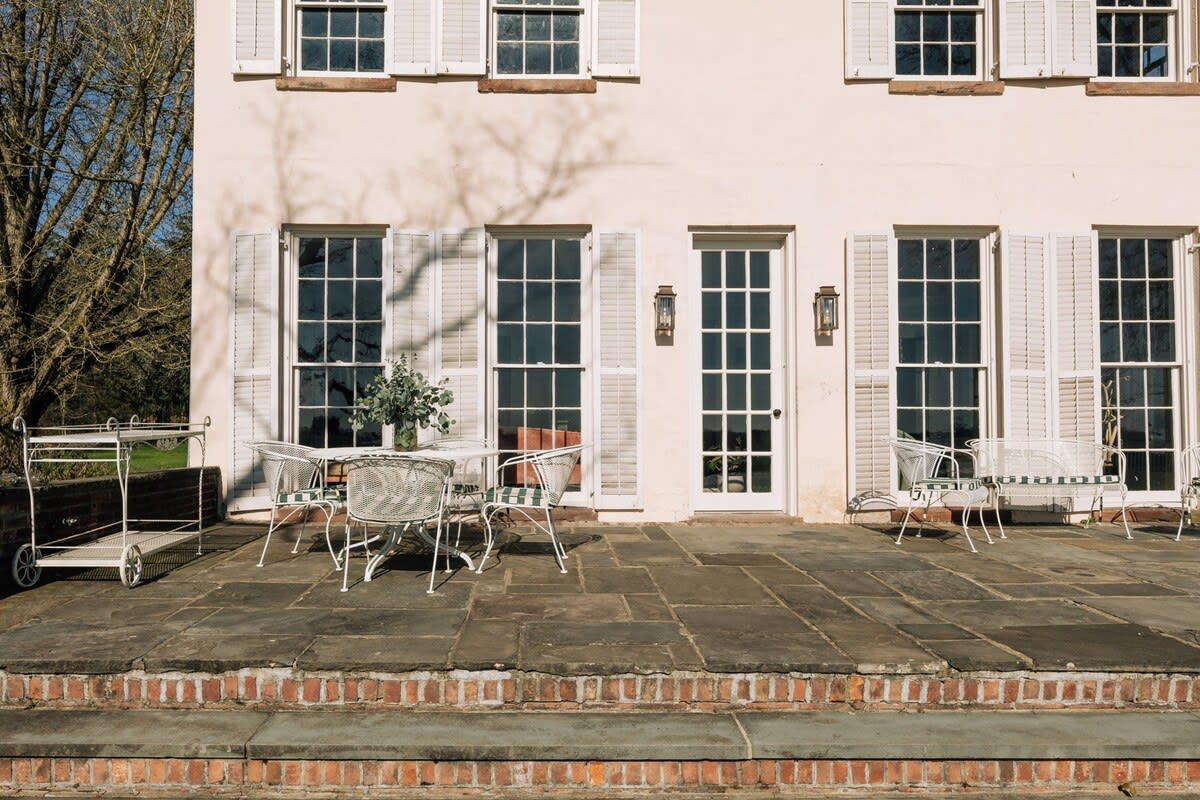 An inviting stone patio furnished with white wire outdoor seating and a table, positioned outside the lighter-colored section of the house with tall windows and white shutters.