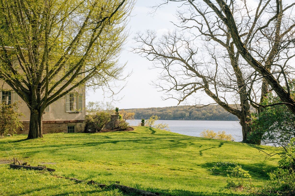 A lush green lawn slopes down towards a wide body of water, with a light-colored house partially visible on the left and mature trees framing the view. The scene is bathed in bright sunlight.