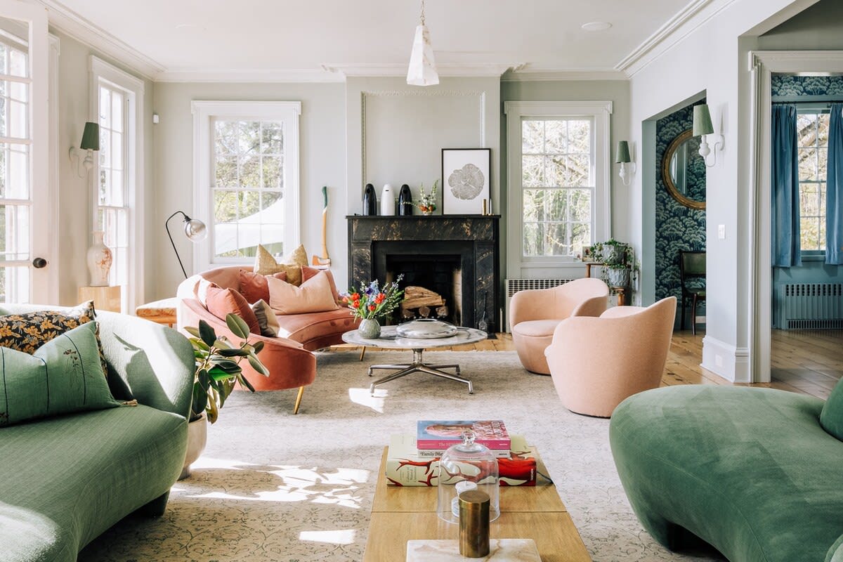 Elegant living room with light green walls, a black fireplace, and a mix of green and pink seating arrangements. Large windows provide ample natural light and views of the outdoors.