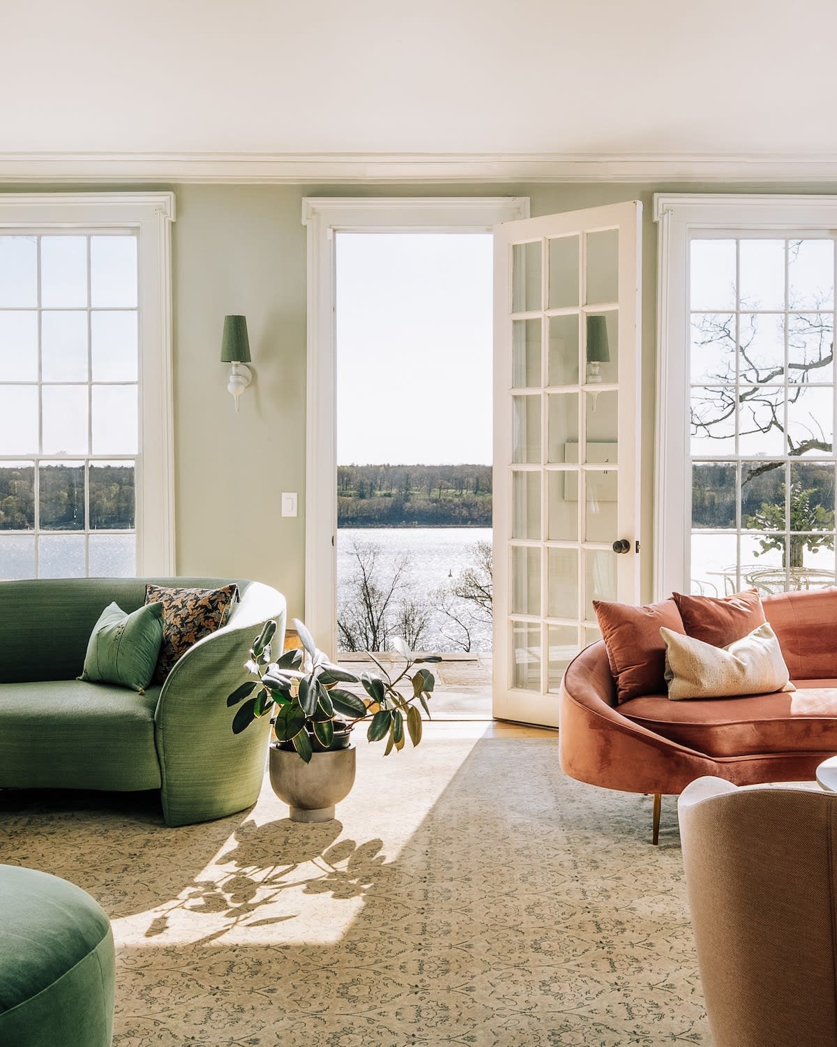 A bright living room with light green walls, featuring a curved green sofa and a curved coral sofa, both facing large windows that offer a scenic view of a lake or river. Sunlight streams through the windows, casting patterns on the patterned rug.