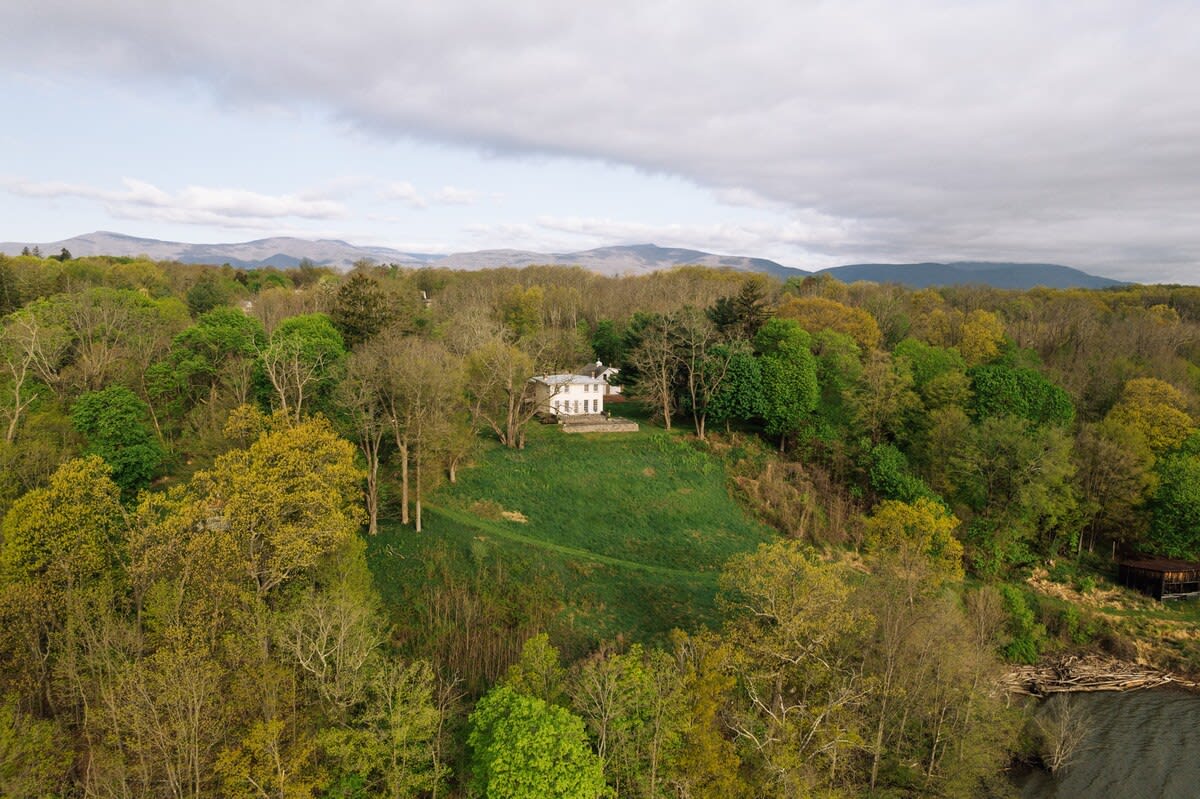 Broad aerial perspective of the white main house situated on a grassy hill, enveloped by vast woodlands with varying shades of green, and distant mountains under a cloudy sky.