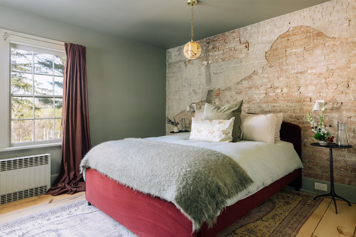 A cozy bedroom featuring a red upholstered bed with a white duvet and a fuzzy grey throw, set against a sage green wall and an exposed brick accent wall. A large window with dark red curtains offers natural light.