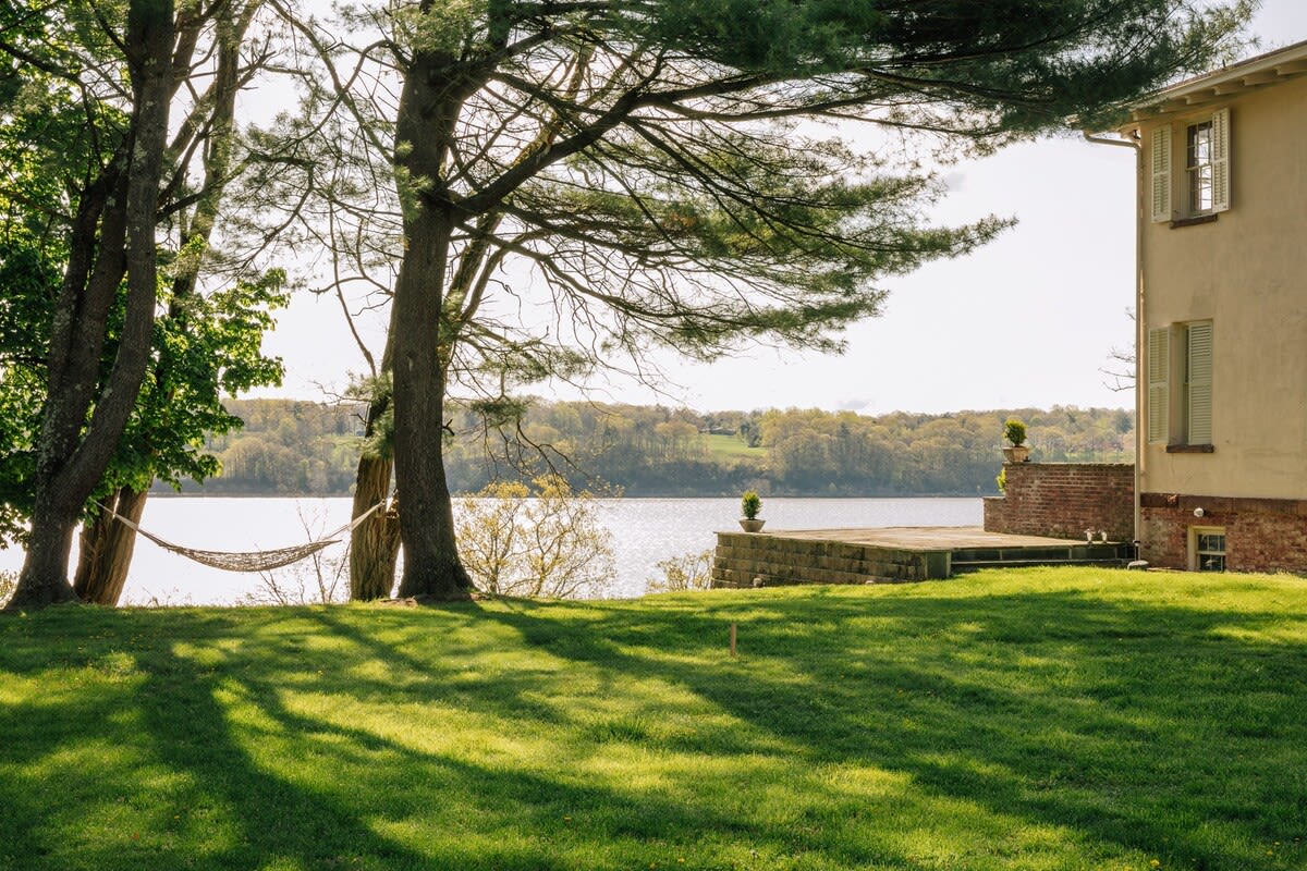 A sunny backyard with a green lawn and mature trees, one of which holds a hammock, leading down to a stone patio overlooking a wide body of water. The side of a light-colored house is visible on the right.