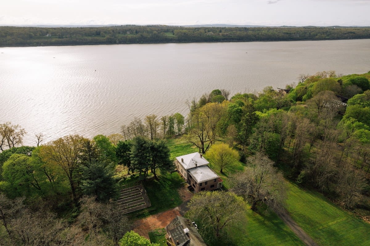 Aerial view of a large property nestled among lush green trees, situated directly beside a wide river or lake under a cloudy sky. The main house has a gray roof.