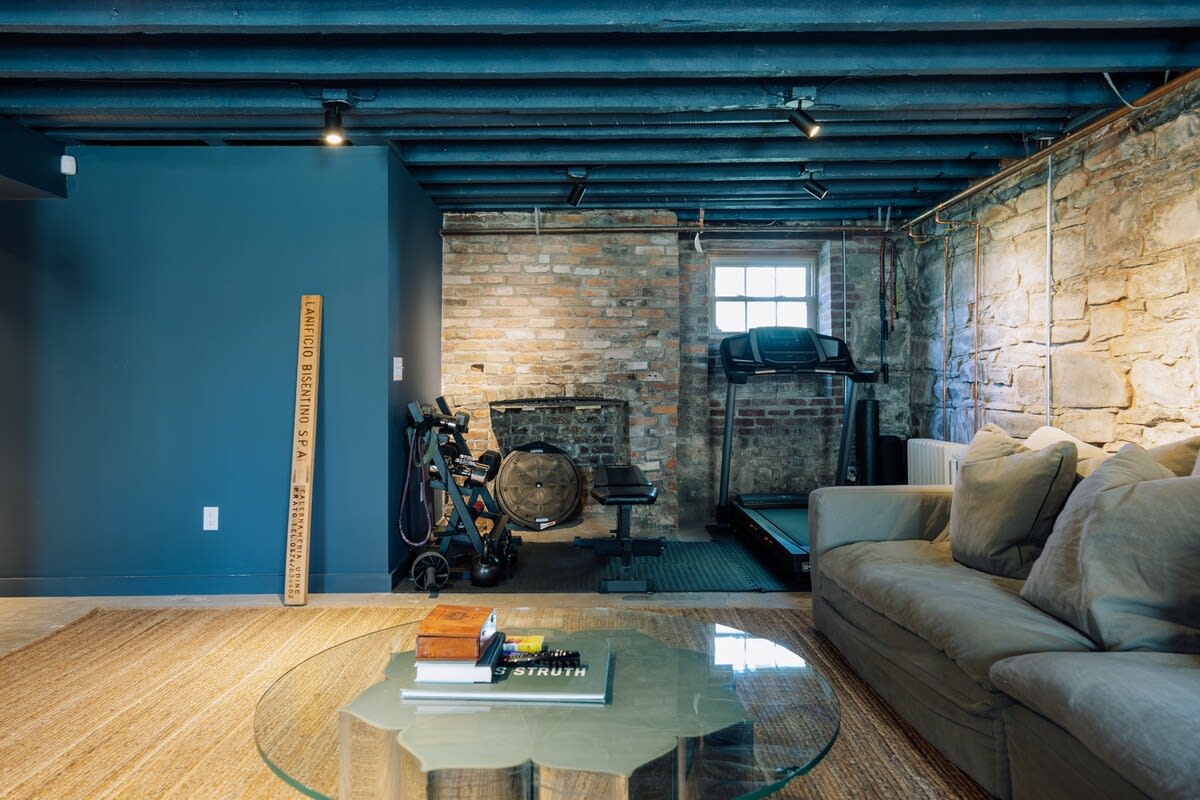 A section of the basement recreation area, continuing the exposed stone and brick walls and black beamed ceiling, featuring a treadmill, weight bench, and other exercise equipment. A grey sectional sofa and glass coffee table are also visible.