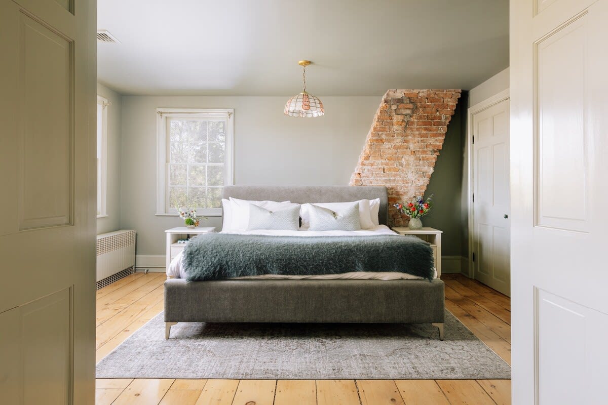 View into a spacious bedroom with a grey upholstered bed, white bedding, and a blue throw, an exposed brick wall, and multiple windows, seen through an open doorway.