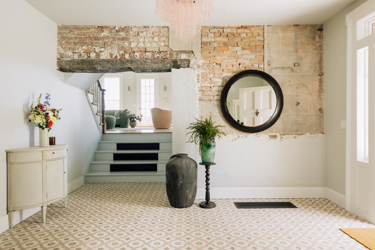 A grand foyer with patterned tile flooring and exposed brick walls, featuring a white staircase leading to an upper level, and a round black mirror above a console table. A large plant in a dark vase adds a touch of greenery.