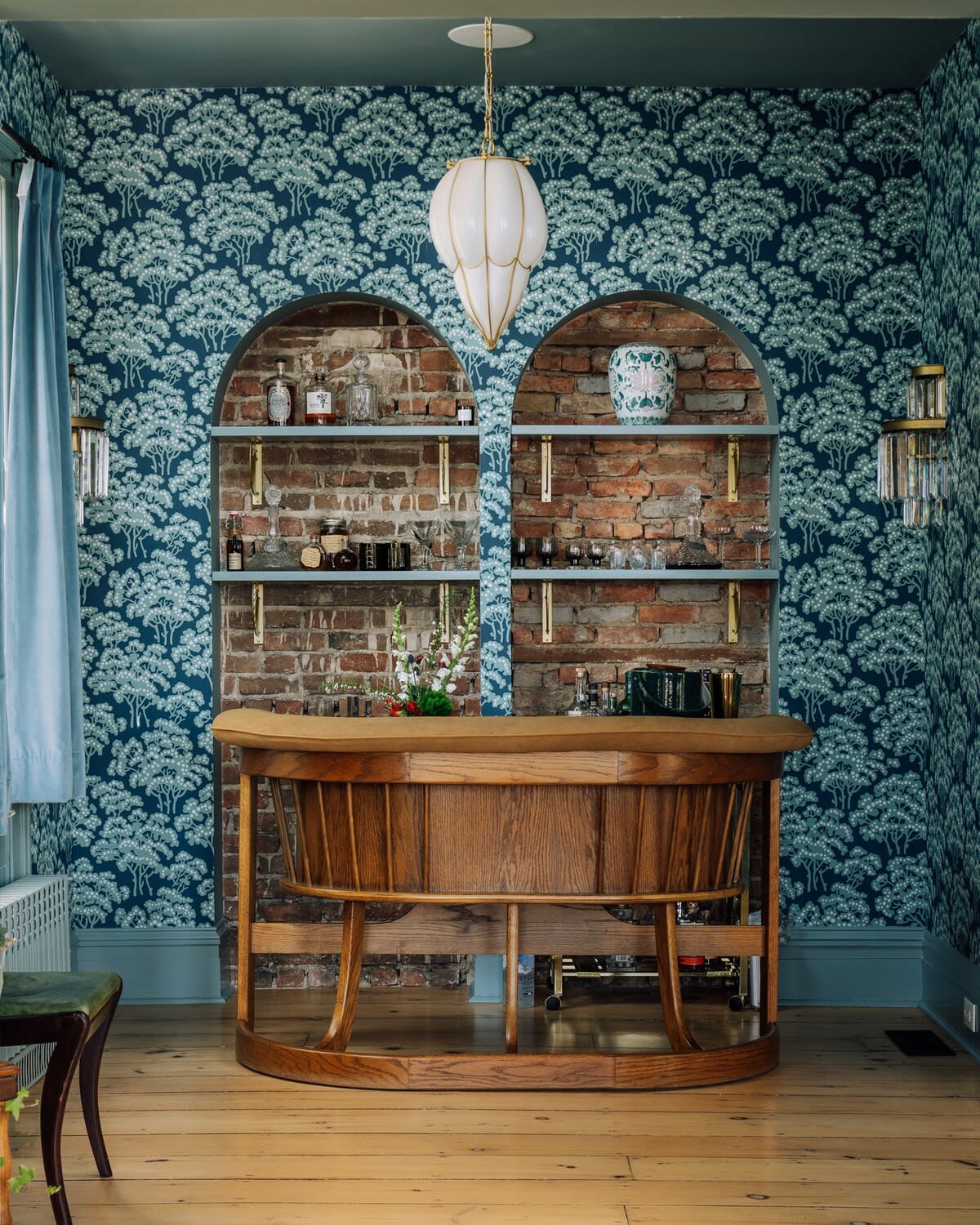 A cozy bar area with dark blue patterned wallpaper featuring tree motifs, exposed brick arched shelves displaying bottles and glassware, and a curved wooden bar counter. A unique pendant light hangs from the ceiling.