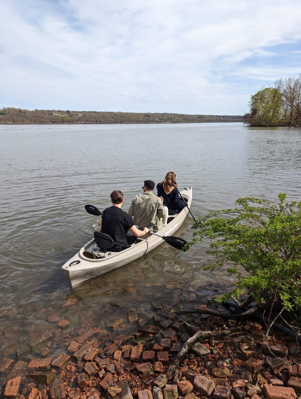 Three people in a tandem kayak paddle on a calm, wide river or lake, with lush green trees lining the distant shore. The foreground shows a rocky bank.