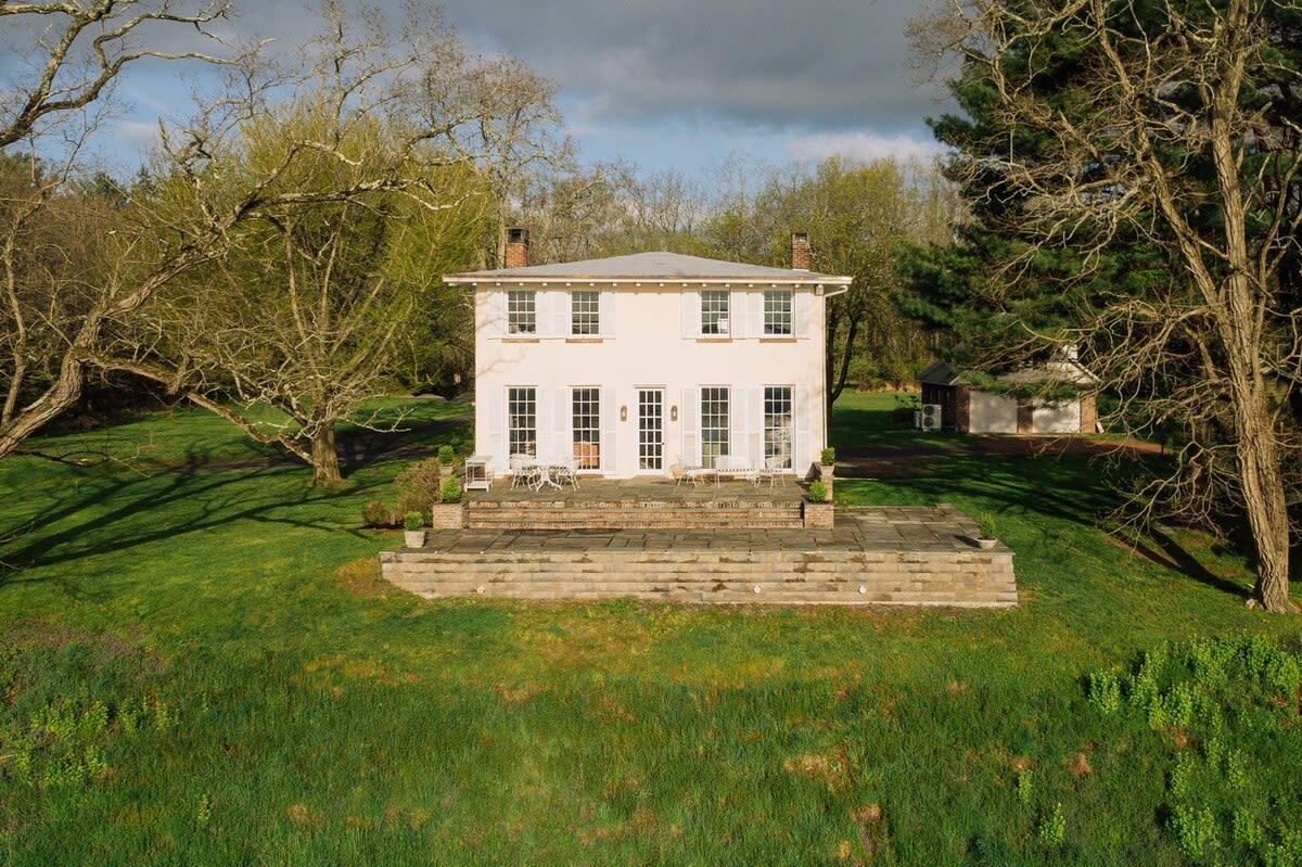 Aerial view of a white two-story house with dark shutters, centered on a large stone patio with outdoor seating, surrounded by a sprawling green lawn and mature trees under a partly cloudy sky.