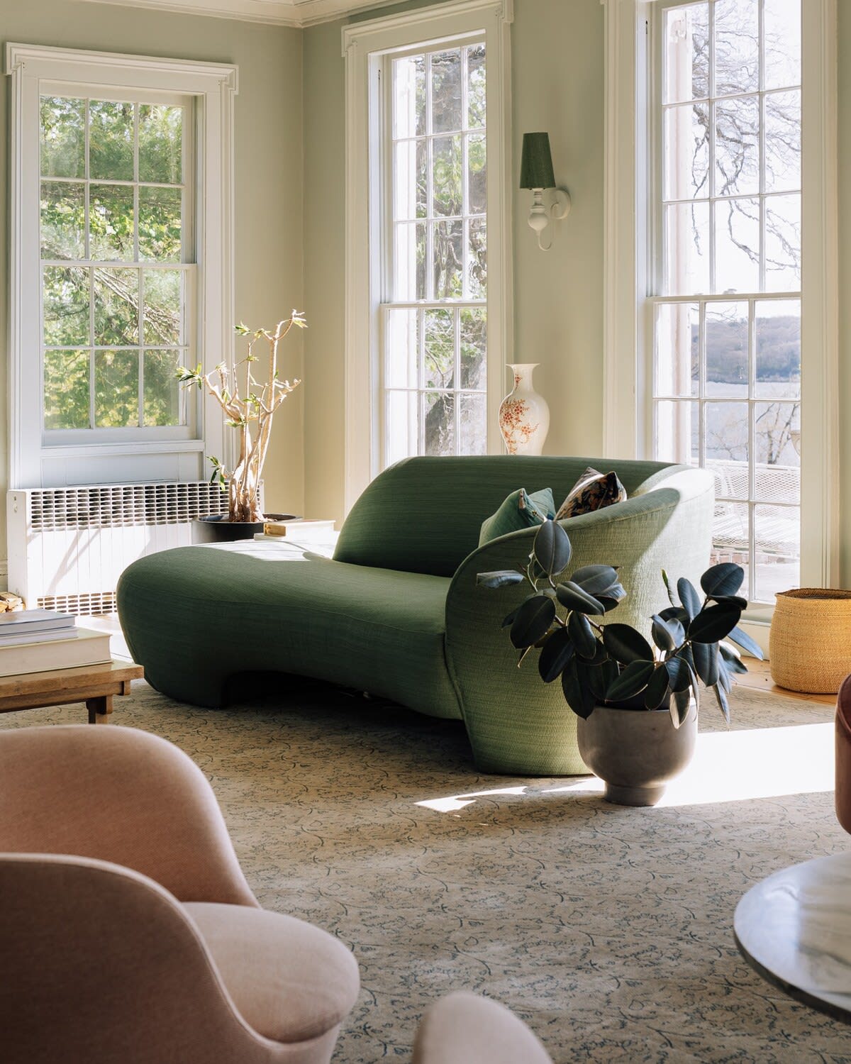 A sunlit living room corner with light green walls and large windows overlooking a lake, featuring a curved green sofa, a light pink armchair, and a dark potted plant. A patterned rug covers the floor, and a white radiator is visible under the window.