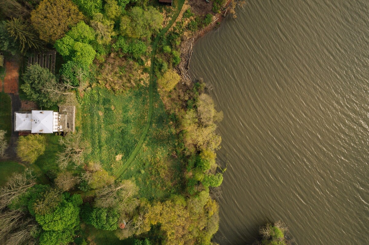 Overhead aerial shot revealing the main house with a light-colored roof, a large manicured lawn, and the textured surface of the river flowing directly adjacent to the property.
