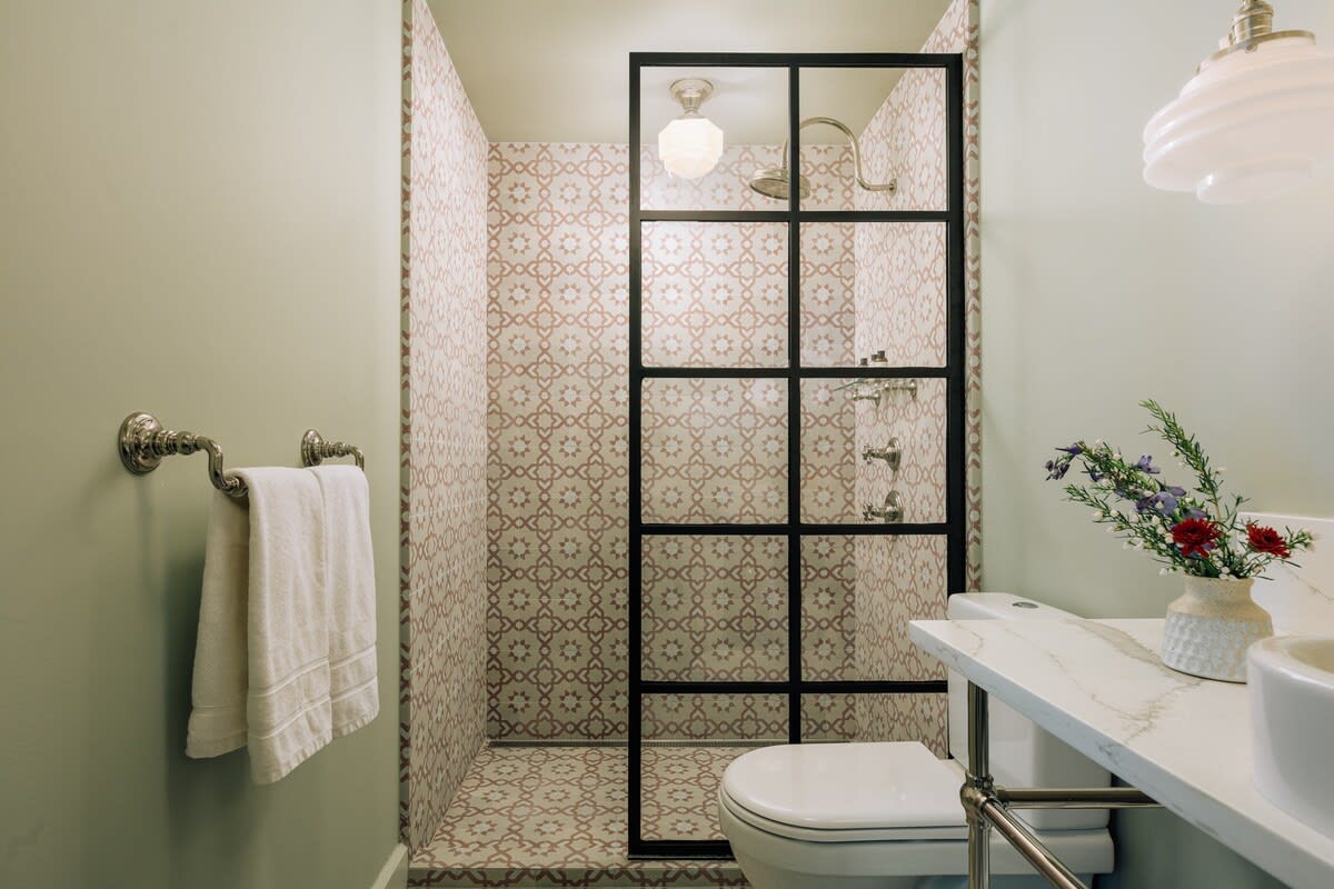 A modern bathroom with light green walls and a walk-in shower featuring patterned geometric tiles in pink and white, enclosed by a black-framed glass door. A white toilet and a vanity with a white countertop are also visible.