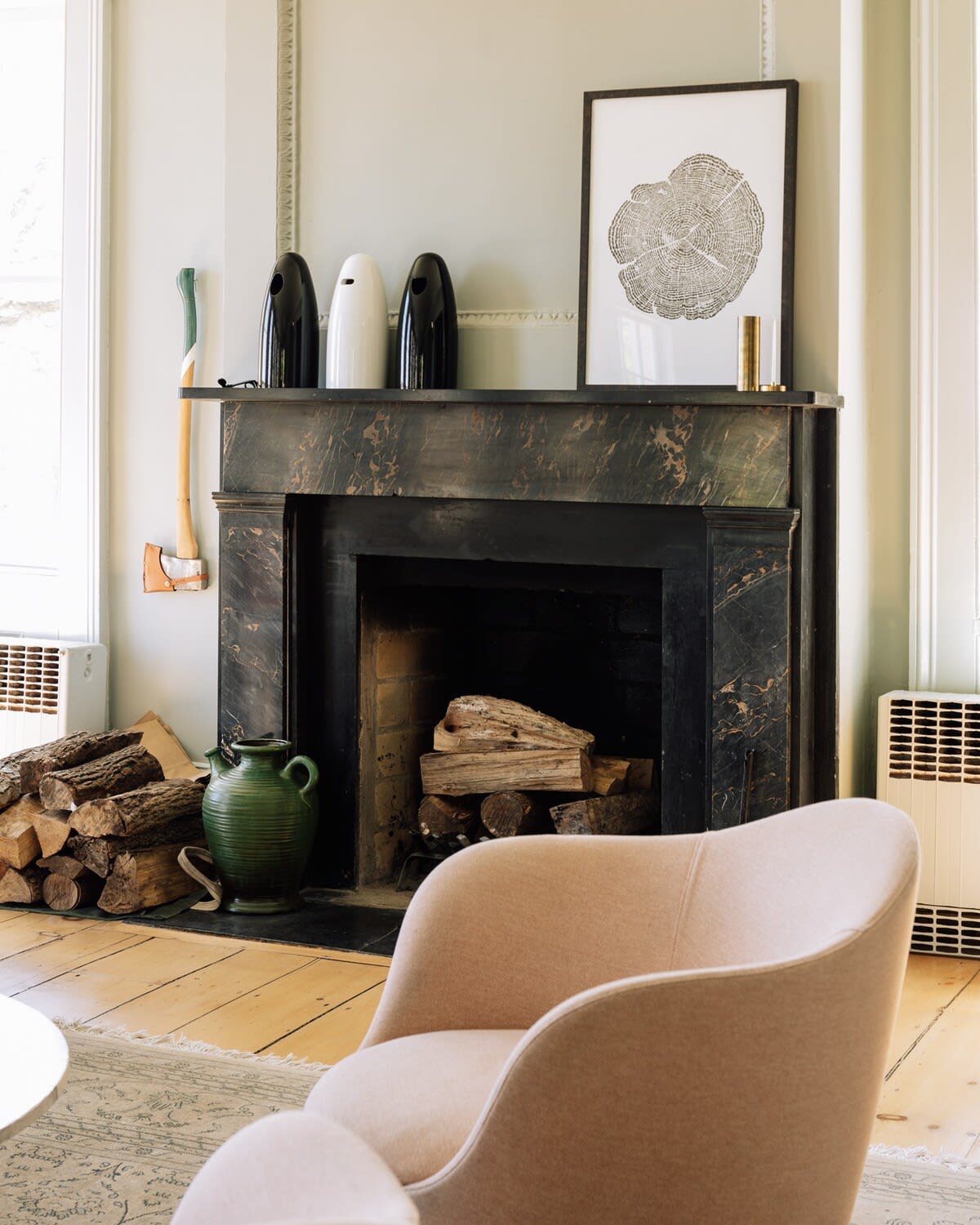 A detailed view of a black marble fireplace with stacked firewood, flanked by a green ceramic vase and an axe, in a living room with light walls. A framed tree ring print hangs above the mantel, and a light pink armchair is visible in the foreground.