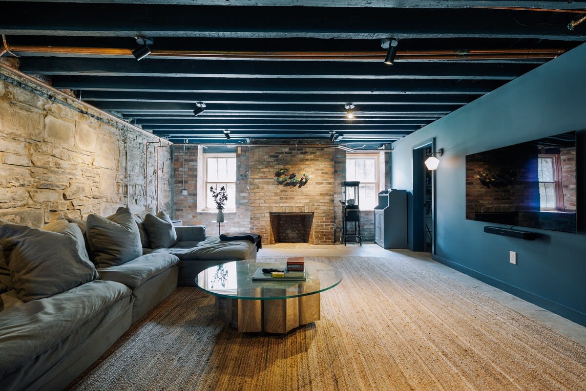 A spacious living area in a renovated basement featuring a large grey sectional sofa, a glass coffee table, and exposed stone and brick walls. The ceiling has exposed black beams with track lighting, and a large TV is mounted on a dark blue accent wall.