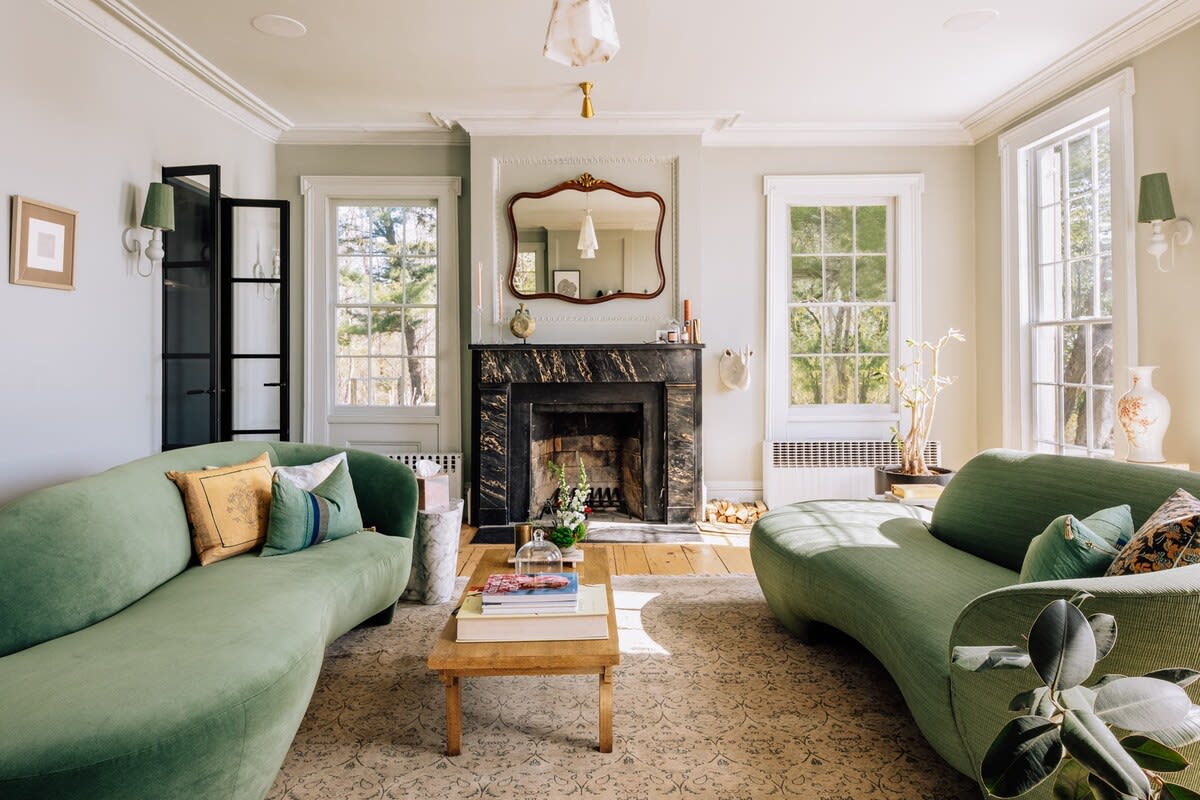 An elegant living room with light green walls, featuring two curved green sofas, a dark marble fireplace with a decorative mirror above, and large windows. A wooden coffee table sits on a patterned rug, and the room is filled with natural light.