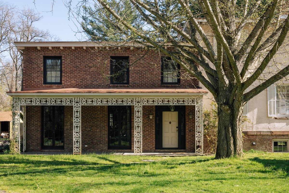 Detailed front view of the main house's brick section, featuring a charming white decorative iron porch with multiple columns and large windows, set against a green lawn.