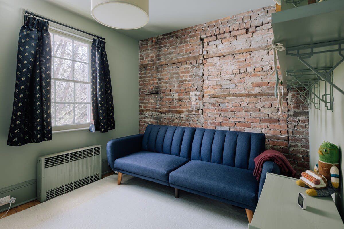 A cozy room with light green walls and an exposed brick accent wall, featuring a blue upholstered sofa, dark patterned curtains, and a window with a radiator below.