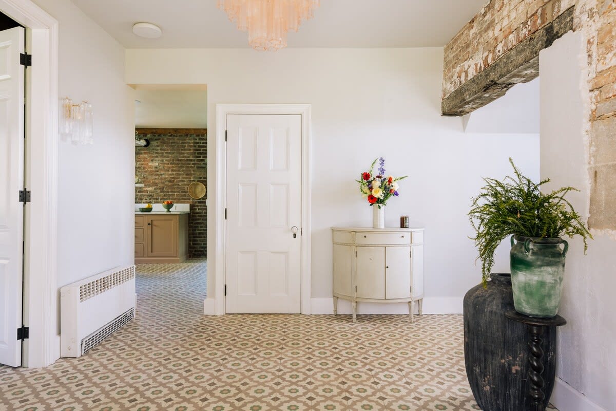 A bright foyer with patterned tile flooring and exposed brick accents, featuring a white console table with a floral arrangement, and a white door leading to another room. A large dark vase with greenery stands on a pedestal.