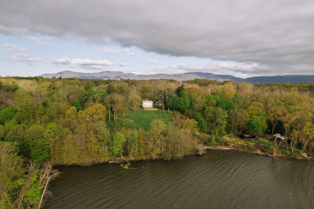 An aerial view showcases a large white house nestled amidst a dense forest of green trees, situated on a hill overlooking a wide body of water. Distant mountains are visible under a cloudy sky.