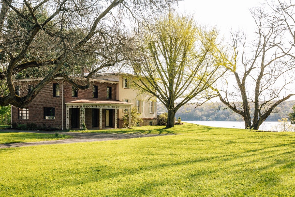 The stately main house, featuring a brick section and a lighter-colored facade, stands behind a wide green lawn, framed by large, bare trees, with the river sparkling in the background.