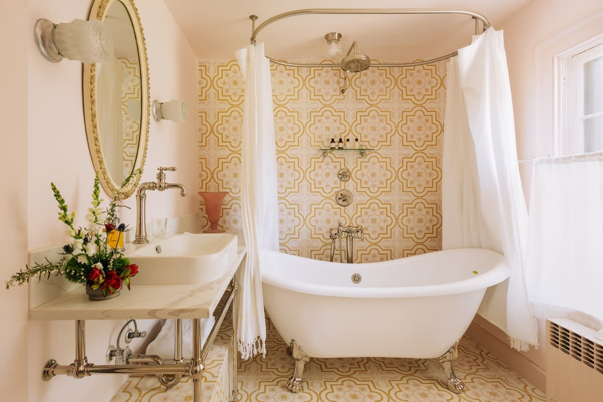 A bright bathroom with light pink walls and patterned yellow and white floor and shower tiles, featuring a classic white clawfoot bathtub with a shower curtain. A white vanity with a vessel sink and an ornate mirror completes the space.