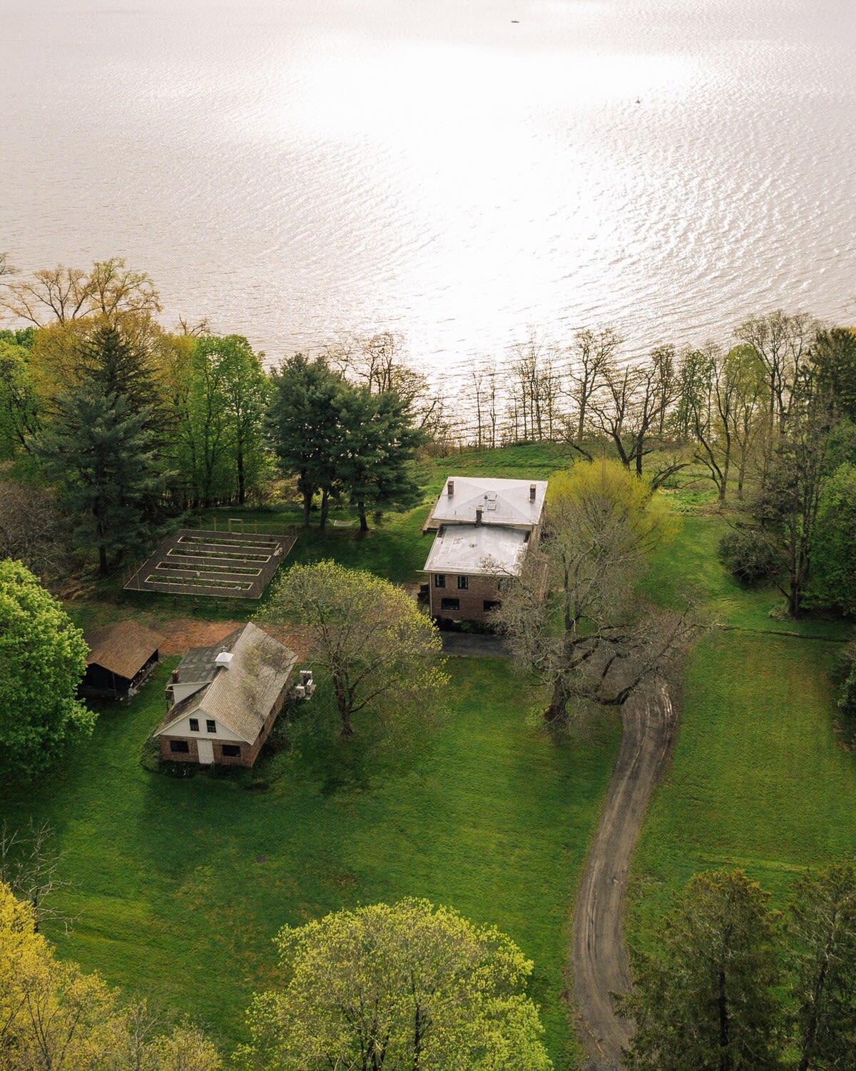 Vertical aerial shot capturing the main house, a separate smaller building, a structured garden plot, a winding dirt path, and the sun-dappled river.
