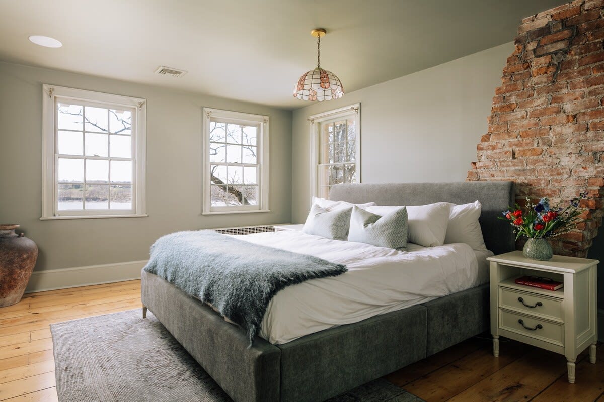 A cozy bedroom featuring a large grey upholstered bed with white bedding and a fuzzy blue throw, an exposed brick wall accent, and two large windows offering views of the outdoors.