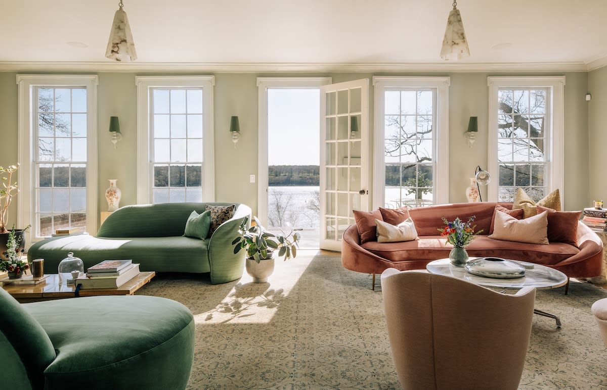 Bright living room with light green walls, two curved sofas (one green, one pink), and large windows offering expansive views of a lake or river. Natural light floods the patterned rug.