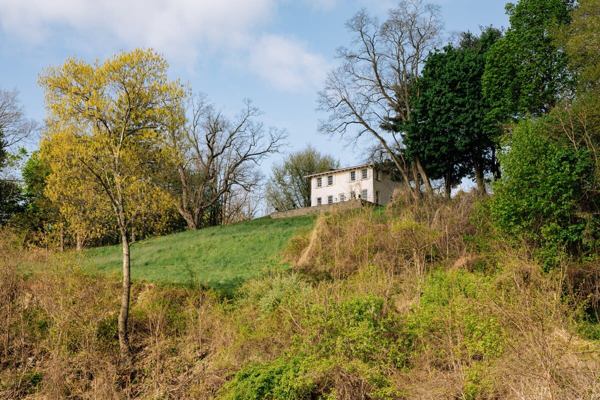 The white-sided main house is perched on a vibrant green hillside, surrounded by a mix of bare and budding trees under a bright blue sky with scattered clouds.