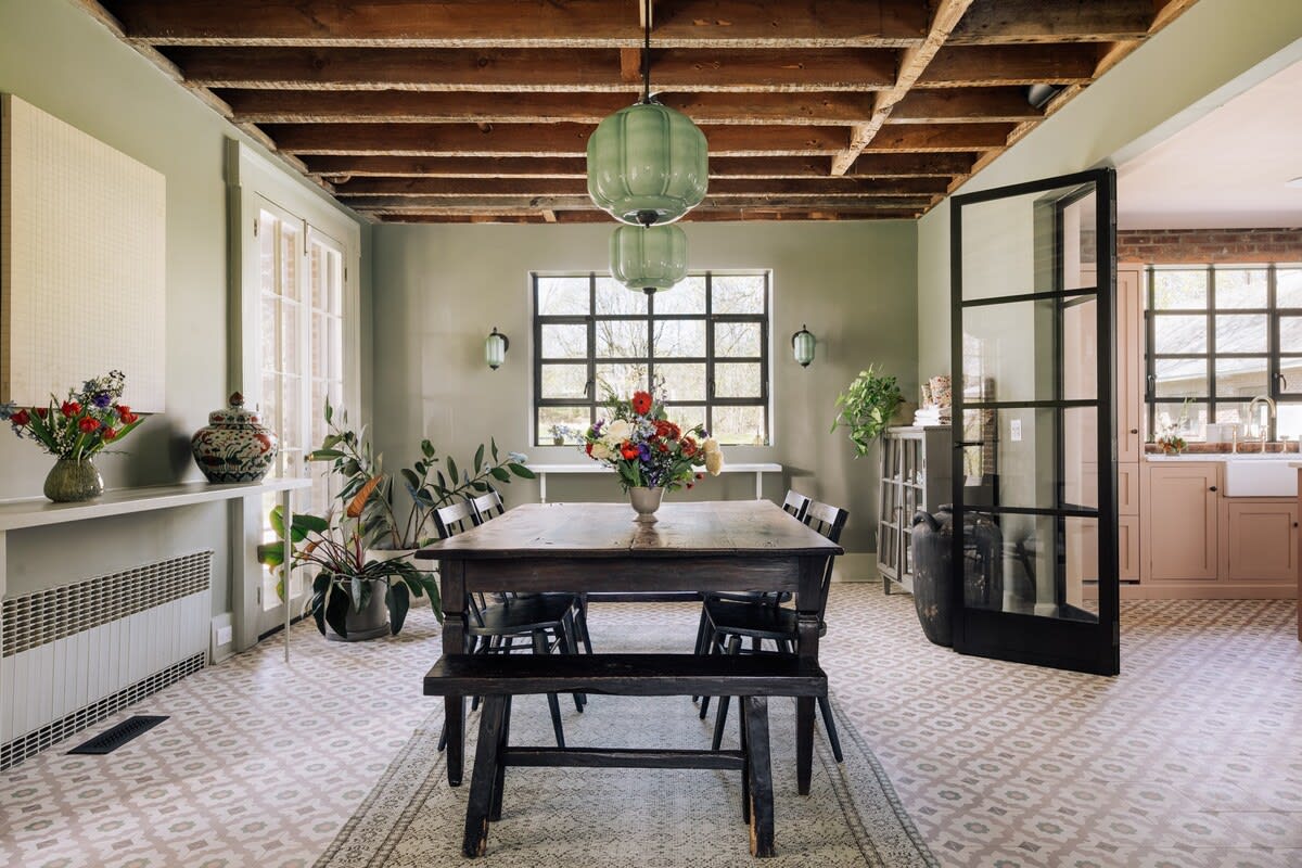 Dining room with an exposed wooden beam ceiling and light green walls, featuring a rustic dark wood dining table with benches and chairs. Large windows and a glass door connect to the kitchen.