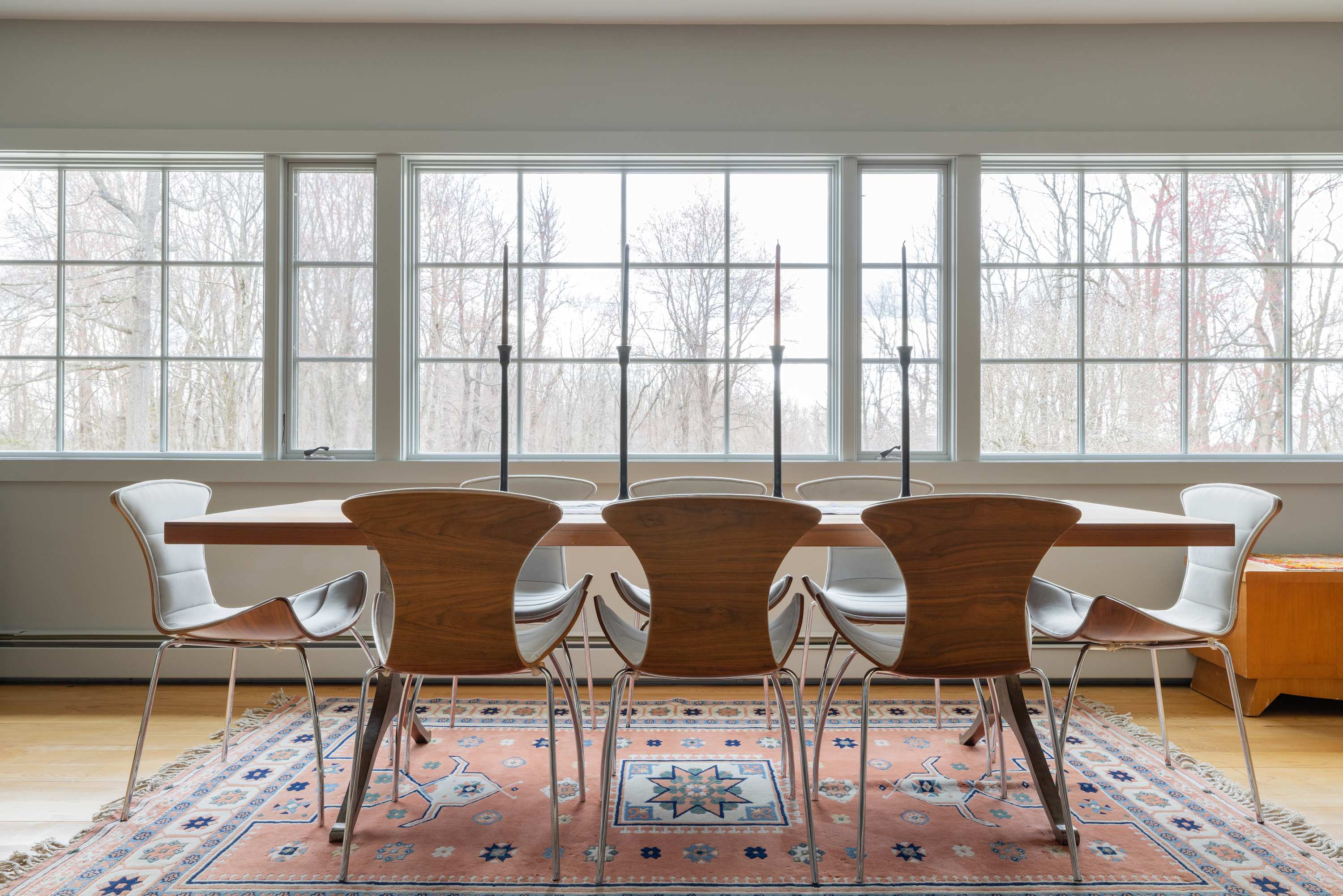 Bright dining area featuring a long wooden table with six modern chairs, set on a pink and blue patterned rug. Large windows overlook a wooded landscape, providing ample natural light.