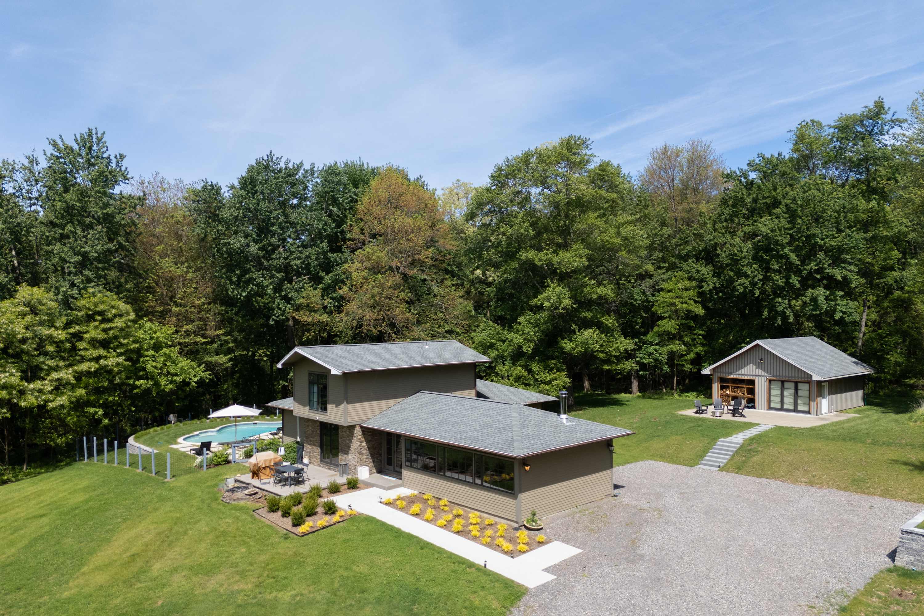 An aerial perspective of the property, highlighting the multi-level main house with a gray roof and a separate gabled utility building. A swimming pool is visible to the left of the main house, all surrounded by expansive green lawns and mature trees.