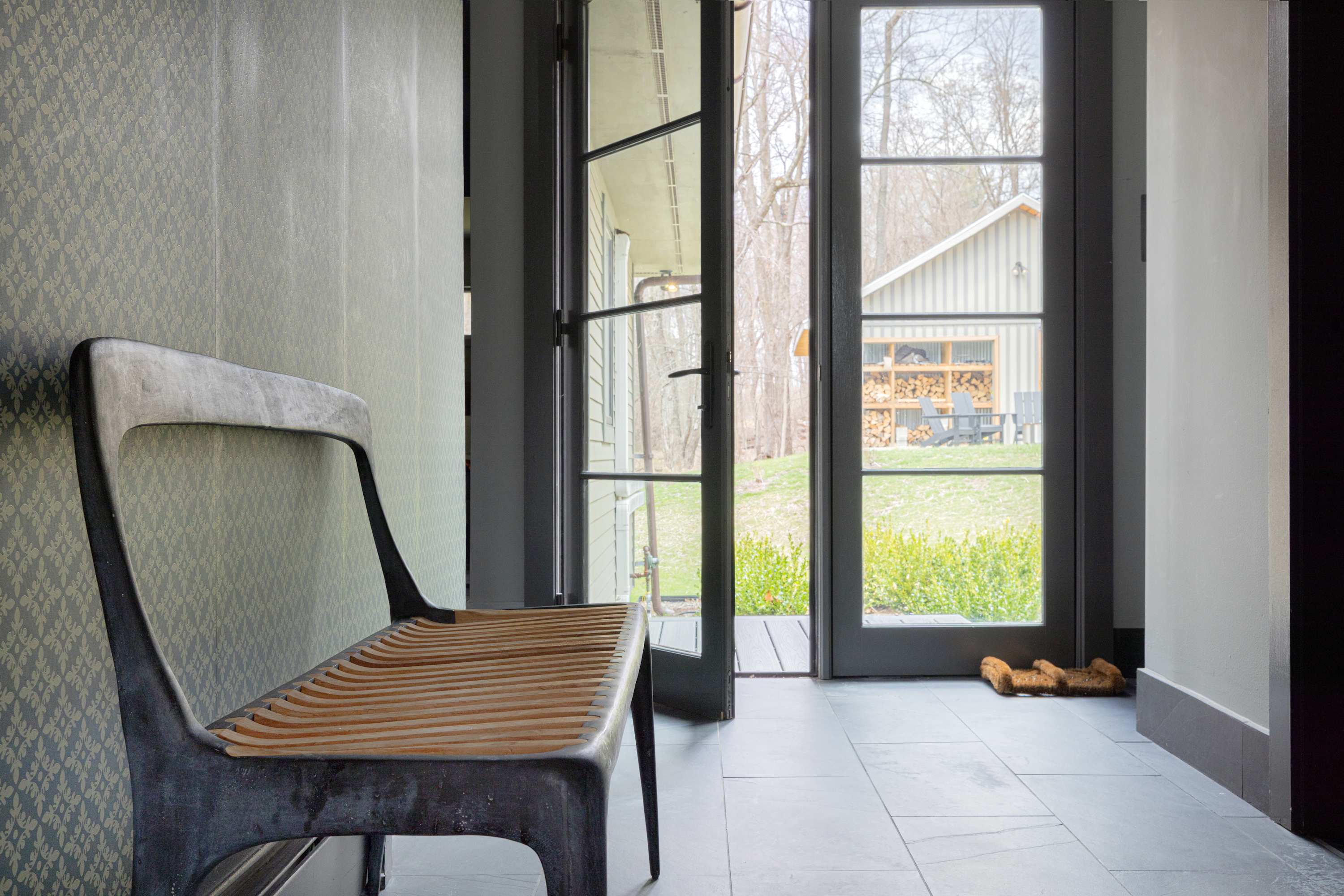 A minimalist foyer with light gray tiled floors and patterned wallpaper, featuring a modern dark metal bench with a wooden seat. Large glass doors open to an outdoor area with a view of a building and trees.
