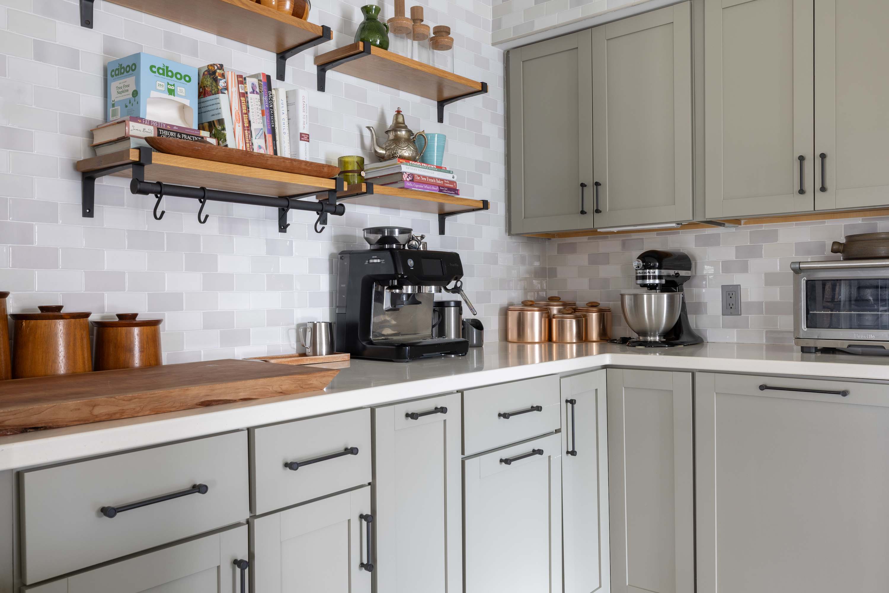 Modern kitchen with light gray subway tile backsplash, sage green cabinets, open wooden shelving, and stainless steel appliances including a coffee maker and stand mixer. Copper accents add warmth to the space.