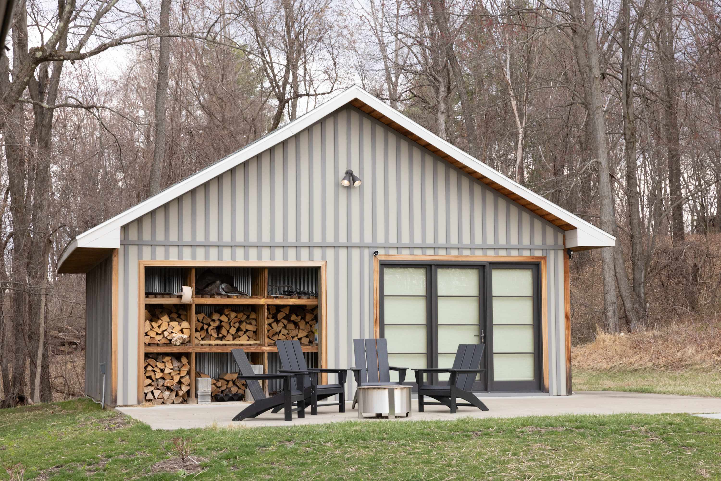 A modern gray utility building with vertical siding and a gabled roof, featuring an open section for firewood storage and a sliding glass door. An outdoor seating area with dark Adirondack chairs and a fire pit is set on a concrete pad.