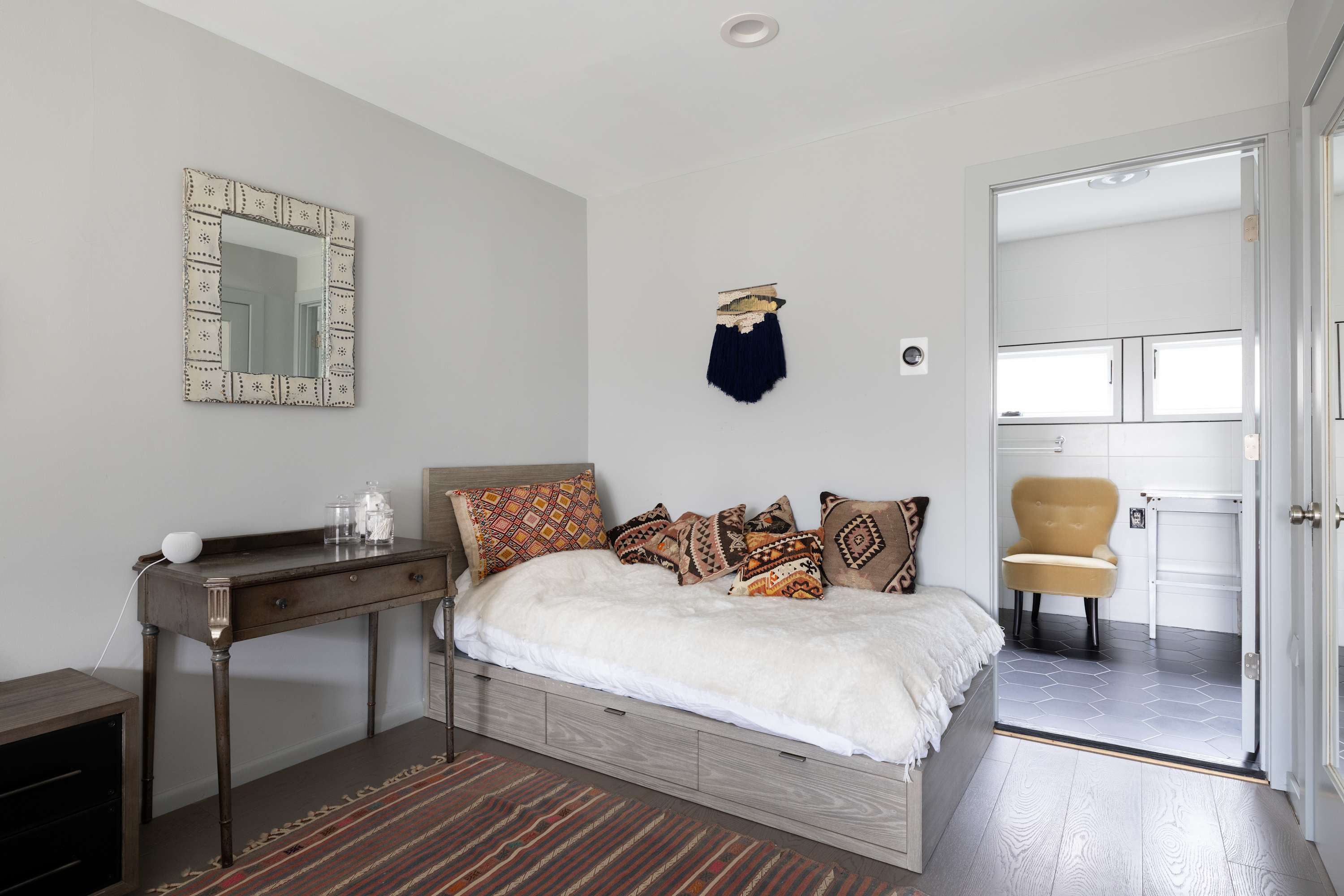 A cozy bedroom featuring a light wood-framed daybed with patterned pillows and a white fluffy blanket. A rustic wooden desk with a decorative mirror is on the left, and an open doorway leads to a bathroom.