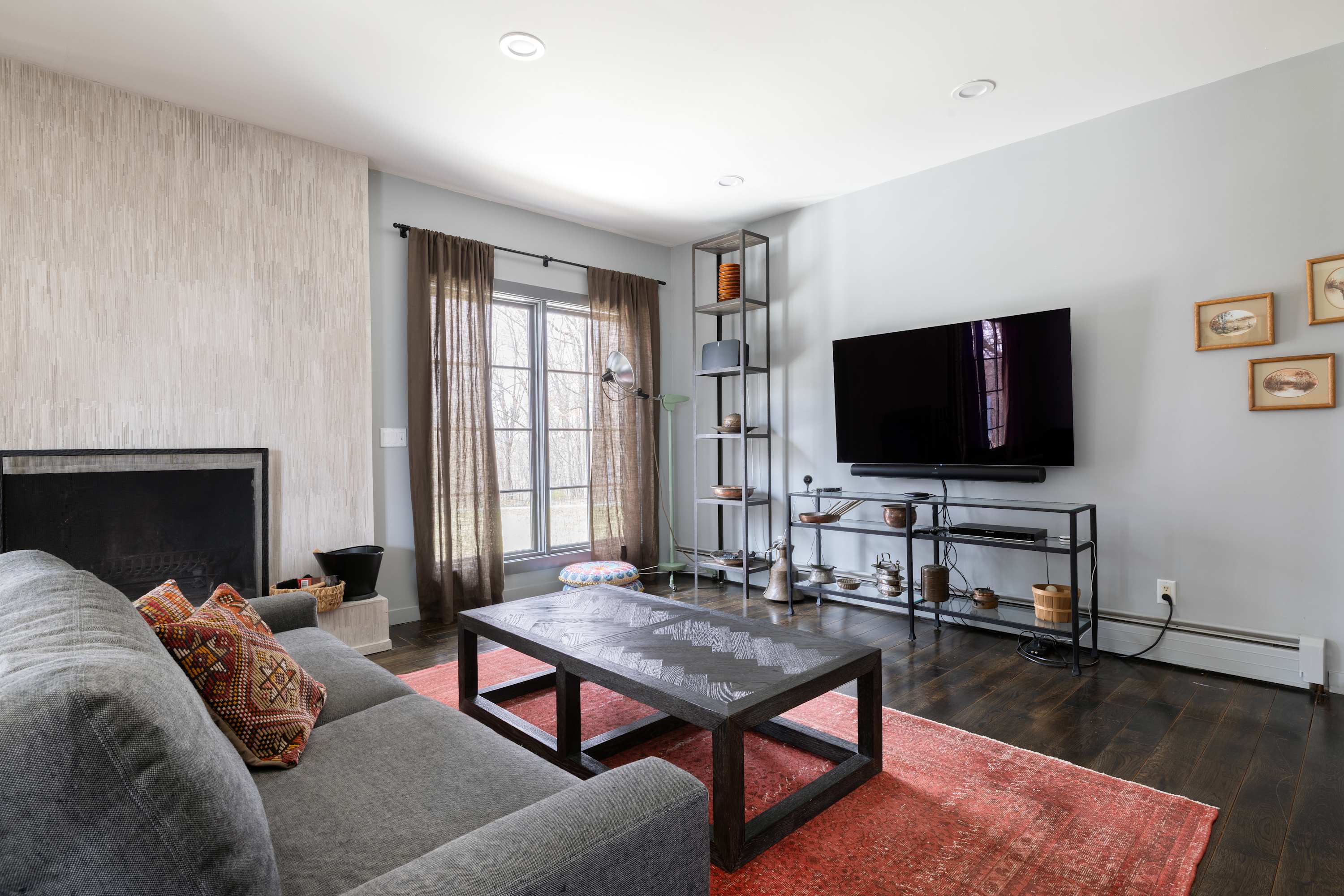 Cozy living room featuring a gray sofa with patterned pillows, a dark wood coffee table, and a large flat-screen TV on a black metal stand. A textured white fireplace and a red patterned rug add character.